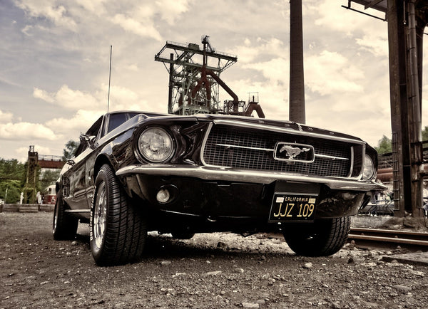 Ретро черен Ford Mustang с калифорнийски регистрационен номер, паркиран на индустриален терен с метални конструкции и комини на заден план, заснет от нисък ъгъл.