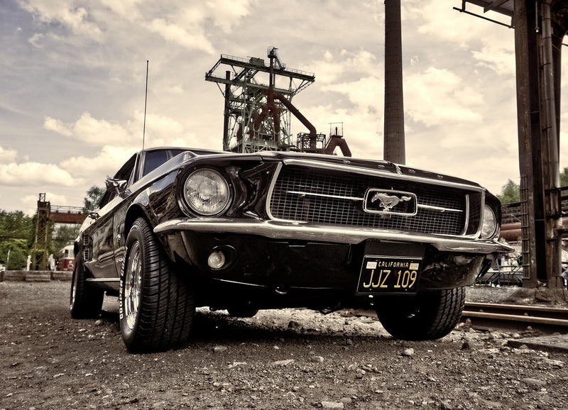 Ретро черен Ford Mustang с калифорнийски регистрационен номер, паркиран на индустриален терен с метални конструкции и комини на заден план, заснет от нисък ъгъл.